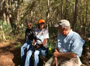 Three community members sitting on a bench in the preserve surrounded by trees. 